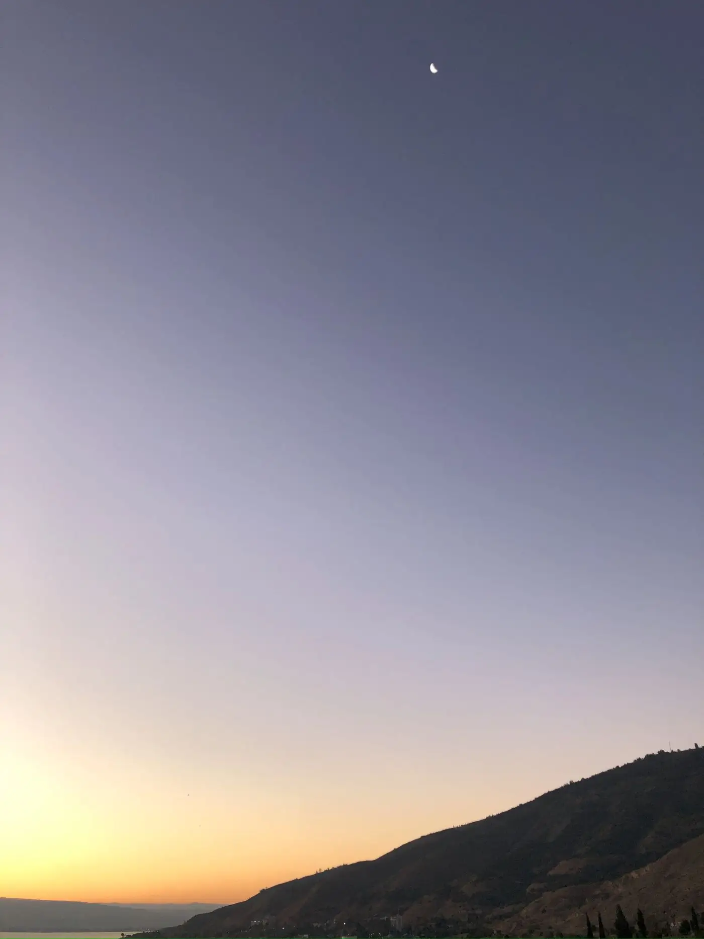 A crescent moon over a hillside at dusk, calm water below.